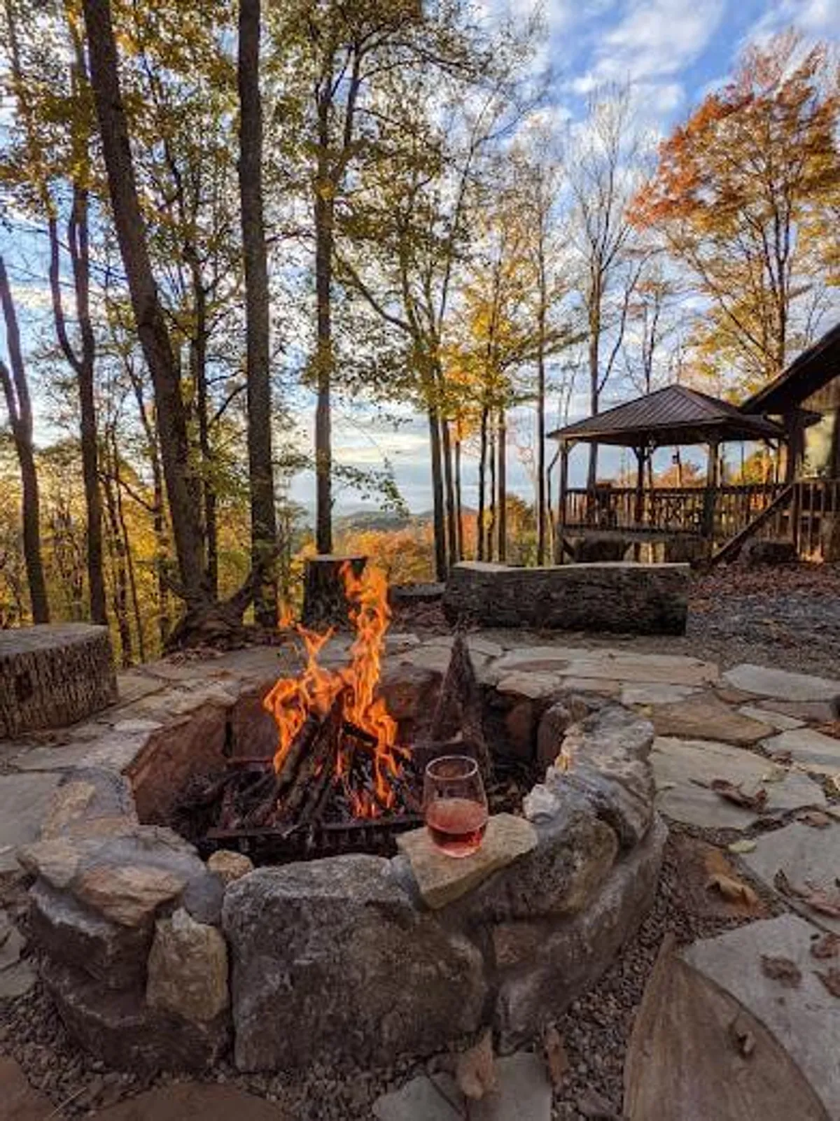 This image captures an exterior fire pit surrounded by cozy seating, with flames flickering as a glass of wine rests nearby. Enveloped by trees and a gazebo, the setting glows in the warm hues of sunset, framing the Blue Ridge Mountains in the distance.