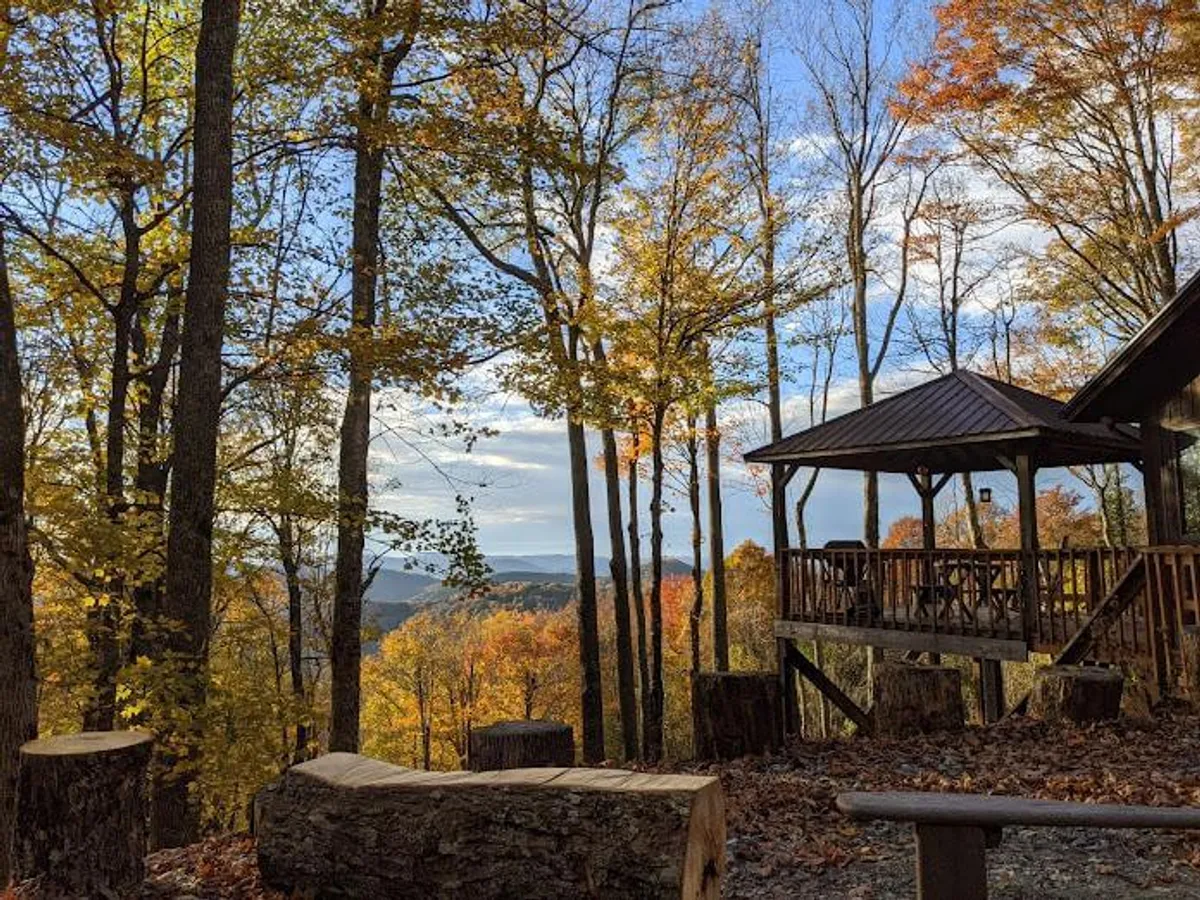 A wooden gazebo sits amongst vibrant autumn foliage, offering a breathtaking mountain view. Logs rest on the ground nearby, suggesting a space to relax before or after a hike. The scenery is awash with the warm colors of fall, enhancing the natural beauty of the surrounding area.