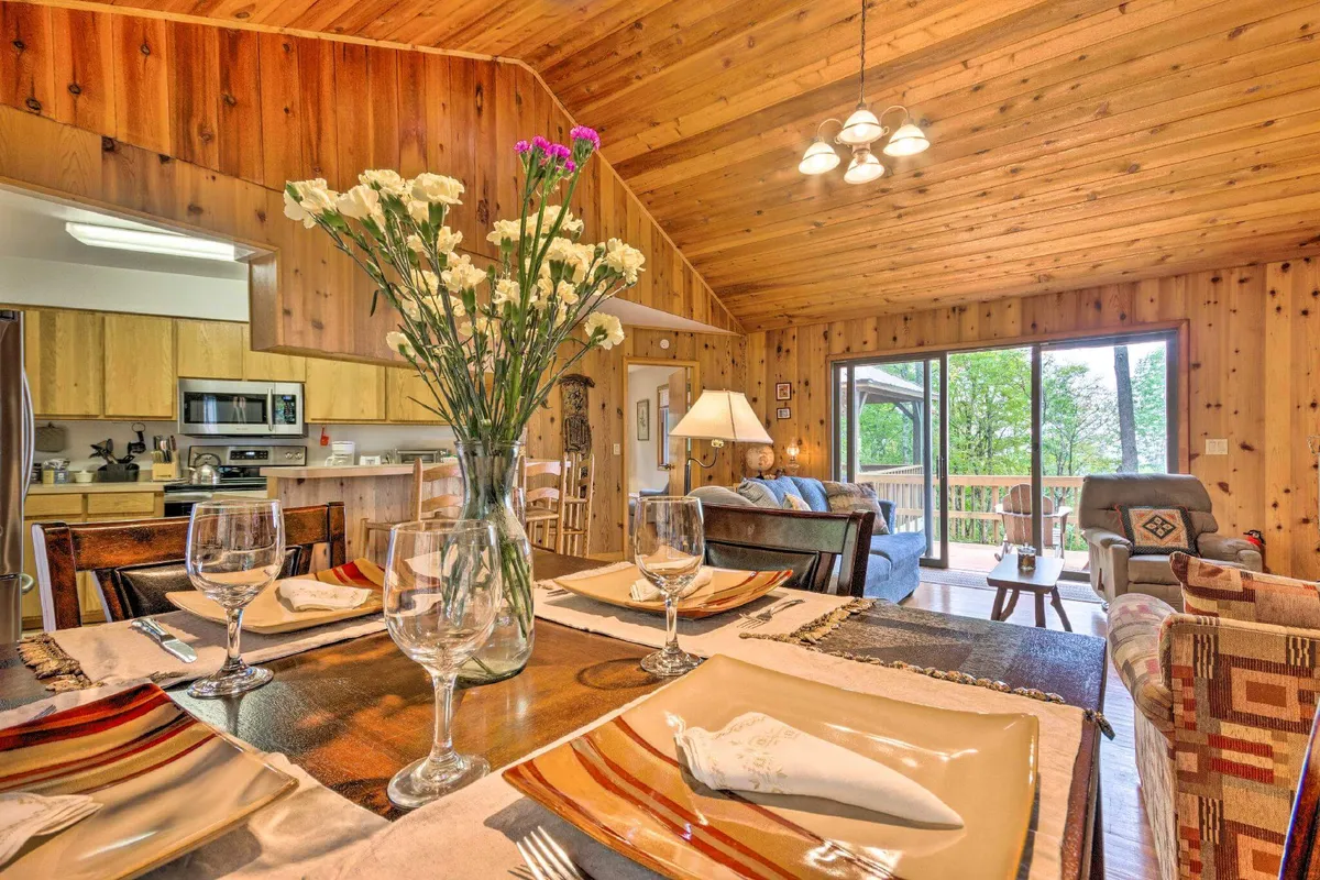 A bright, cozy area with a dining table set for a meal, floral centerpiece, and a comfortable seating nook by large windows that let in plentiful natural light and offer glimpses of the outdoors, all surrounded by warm wooden details.