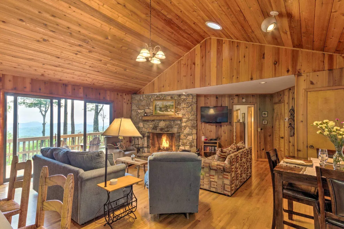 A spacious living room with a central stone fireplace, large windows framing expansive mountain views, and a comfortable seating area arranged for relaxation or group gatherings. The room also features a dining table, blending social and dining spaces under warm, natural light.